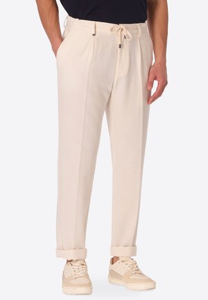 Uomo con pantaloni color crema con coulisse e orli risvoltati e sneakers crema, mani in tasca, su sfondo semplice.