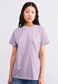 Camiseta de lavanda clara con cuello redondo, mangas cortas y un logo de marca blanco en el cuello, fabricada en suave tela de algodón.
