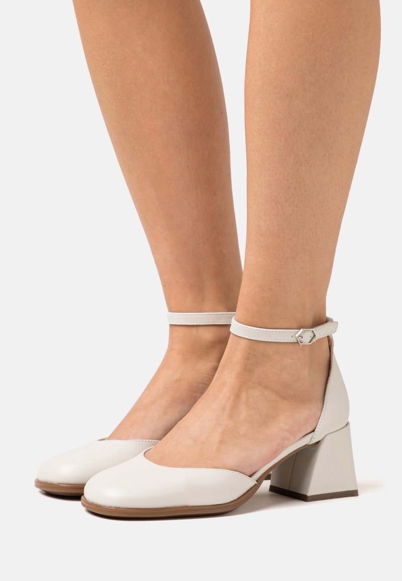 Chaussures blanches en cuir à bout fermé, talon épais, avec brides à la cheville et boucles argentées, portées pieds nus sur un fond uni.