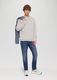 Hellgrauer strukturierter Pullover, blau kariertes Hemd über die Schulter gehängt, blaue Jeans und weiße/graue Sneakers. Einfaches, lässiges Outfit.