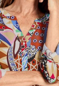 Bunt gemusterte Bluse mit V-Ausschnitt, Knopfverschluss und floralen/grafischen Druckmotiven. Der Stoff hat eine glatte Textur.