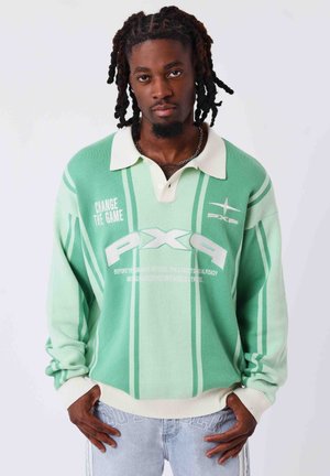 Homme avec des dreadlocks portant un pull vert à rayures avec col blanc et texte, debout les mains dans les poches d'un jean bleu clair.