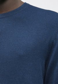 Κοντινό πλάνο ενός ατόμου που φοράει ένα μπλε πλεκτό πουλόβερ με στρογγυλή λαιμόκοψη, δείχνοντας την υφή του υφάσματος και την λαιμόκοψη.