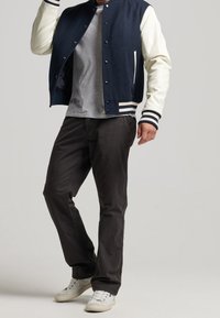 Mörkblå ull varsityjacka med krämfärgade ärmar, randiga manschetter och tryckknappar. Åtsidan med grå t-shirt och svarta byxor, bärande vita sneakers.