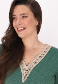 Un top verde cu decolteu în V, decorat cu dantelă bej și detalii intricate, completat de păr natural și accesorii subtile.