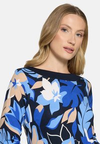 Top s květinovým vzorem v živých modrých, bílých a béžových odstínech. Vyrobený z měkké látky s výstřihem do člunu a dlouhými rukávy.