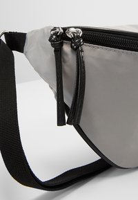 Szara nerka z nylonu z czarnymi akcentami, zakrzywiony kształt, czarny pasek i zamek błyskawiczny z dwoma teksturowanymi uchwytami.