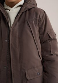 Kadro bližje rjavega podložene jakne z zadrgo in gumbi spredaj, z žepom z manšeto in bež kapuco spodaj.