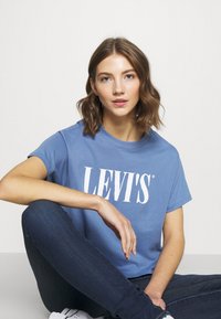Šviesiai mėlyna medvilnės marškinėliai trumpomis rankovėmis su dideliu baltu "LEVI'S®" logotipu priekyje. Susieti su tamsiomis džinsomis.