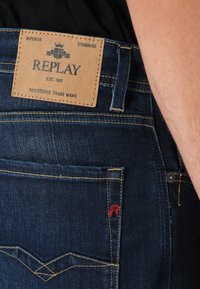 Indigo denimové džínsy s koženou nášivkou s nápisom "REPLAY." Funkcie žltého prešívania a červeného loga na zadnej vrecku, štíhly strih.