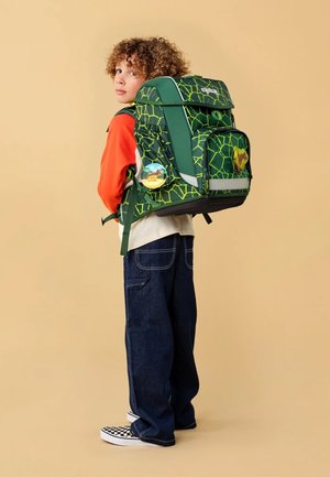 Grüner Rucksack mit hexagonalem Muster, verstellbaren Trägern, mehreren Taschen und einem dekorativen Aufnäher. Das Modell trägt ein rotes Oberteil und Jeans.