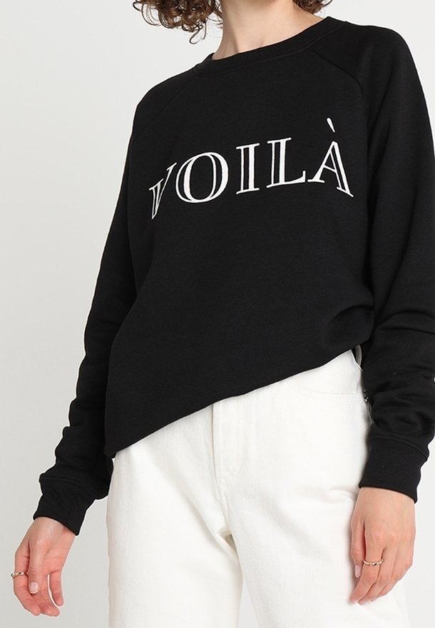 Sweat-shirt noir à manches longues avec le texte "VOILÀ" en blanc, porté avec un pantalon blanc taille haute, présentant une pose décontractée et des bagues aux doigts.