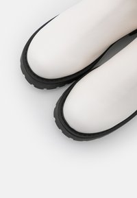 Botas de tornozelo brancas feitas de material suave, com uma sola de borracha preta espessa. O design é aerodinâmico com a ponta arredondada.