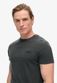 Camiseta de manga corta en gris oscuro con logo bordado en el área del pecho izquierdo. Presenta un cuello redondo y una tela suave y texturizada.