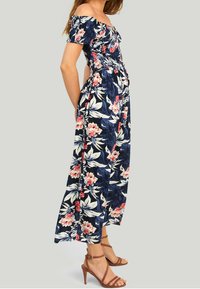 Robe maxi fleurie à design décolleté, en tissu bleu marine, avec des fleurs rouges et blanches, et une taille cintrée. Elle possède des bretelles délicates et des poches.