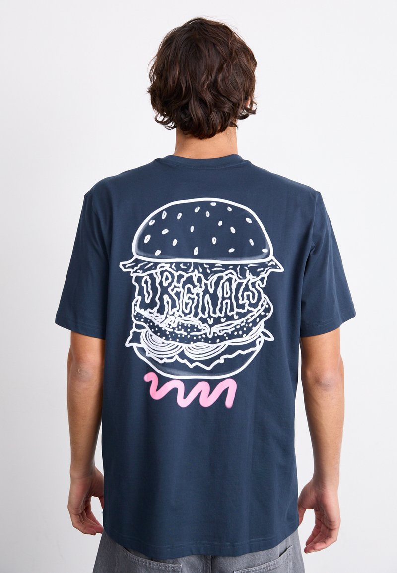 Person som bär en marinblå t-shirt med en stor vit burgargrafik och ordet "ORIGINALS" på ryggen, samt en rosa slingrig linje nedanför.