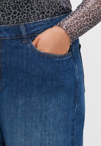 Jeans en denim bleu foncé avec une coupe classique, dotés d'une poche avant et de coutures visibles. Le tissu a une finition lisse et texturée.