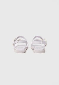 Lilla sandaler med flere stropper, en teksturert såle og en dekorativ blomst detalj på siden. Flatt såle med minimalt med maskinvare.