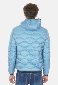 Giacca a vento azzurro chiaro con motivo a onde trapuntate, cappuccio e polsini elastici. Rifinitura nera sul fondo e texture morbida e lucida.