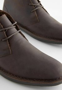 Brązowe skórzane buty typu chukka o gładkiej teksturze, z przeszyciami i dwoma sznurówkami. Podeszwa jest czarna, kontrastująca z górnym materiałem.