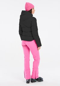 Schwarze Pufferjacke mit Kapuze, rosa Strickmütze und rosa Skihose. Schwarze Skistiefel mit kleinen Reißverschlüssen an den Hosen.