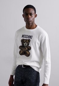Balts garās piedurknes T-krekls ar brūnā lāča grafiku un "Moschino" uzrakstu. Apvienots ar melnajām biksēm, uz kurām redzams Moschino logos.