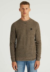 Braun strukturierter Pullover mit Rundhalsausschnitt, gerippten Bündchen und einem kleinen schwarzen Logo-Patch auf der Brust, kombiniert mit blauen Jeans.