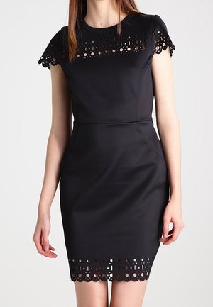 Sort tætsiddende knælang kjole med rund halsudskæring og bølgede laserudskæringer på ærmer, bryst og kant.