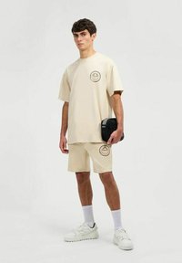 Camiseta beige de manga corta y pantalones cortos a juego, con acentos de logotipo circular. El modelo sostiene un pequeño bolso negro. Zapatos deportivos blancos y calcetines.