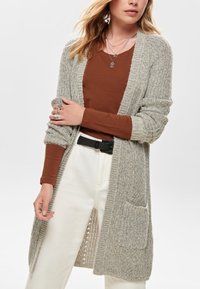 Vrouw draagt een grijze gebreide lange cardigan over een roestkleurig long-sleeve shirt met witte broek en gelaagde colliers.