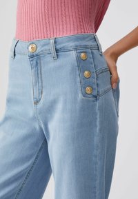 Jean bleu clair taille haute avec trois boutons dorés à la poche latérale, porté avec un haut rose en maille côtelée et une main posée sur la hanche.