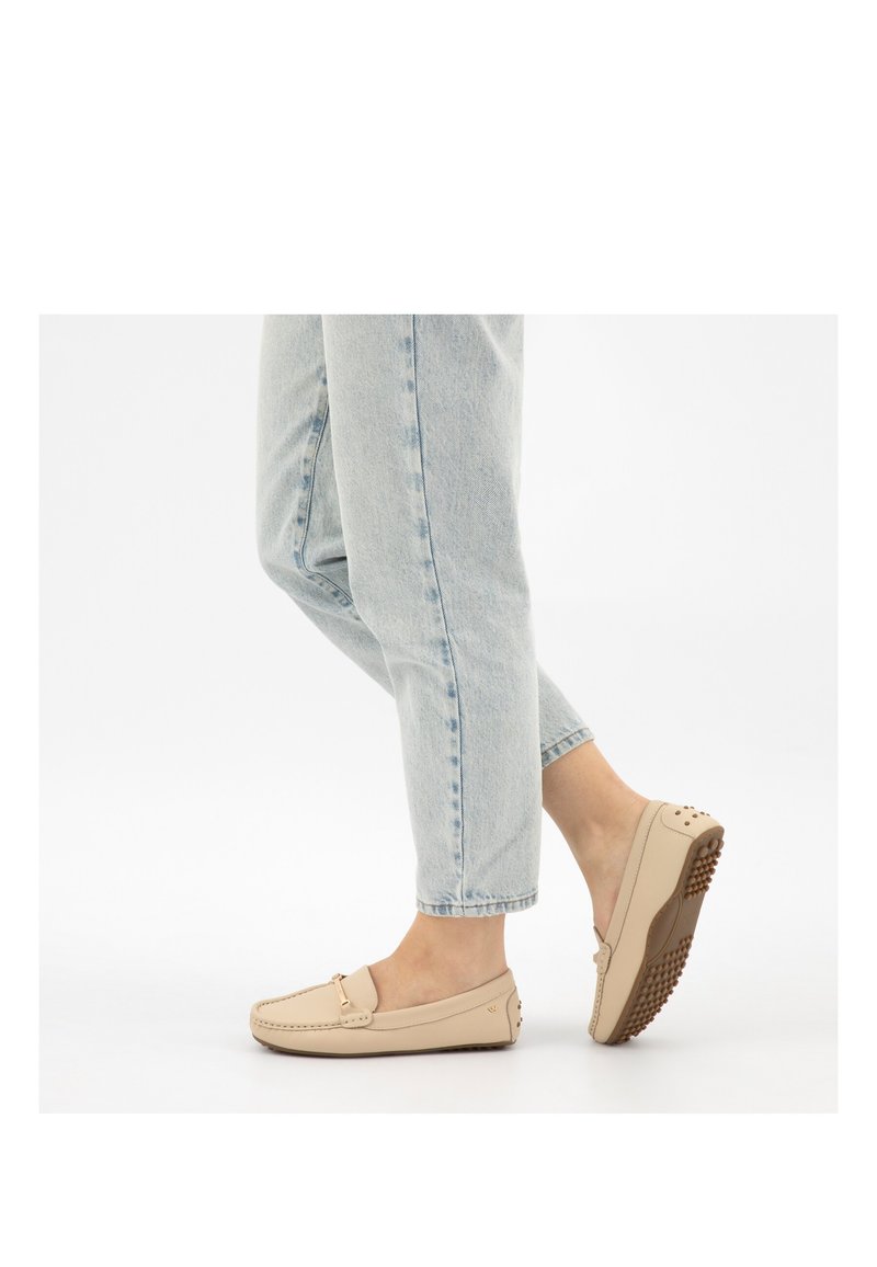 Persoon die lichtblauwe jeans en beige mocassins met rubberen noppenzolen draagt, één voet licht verhoogd, witte achtergrond.