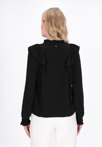 Schwarze Bluse mit langen Ärmeln, Rüschendetails an den Schultern und Manschetten, strukturiertem Stoff und einem kleinen metallischen Detail am hinteren Kragen.