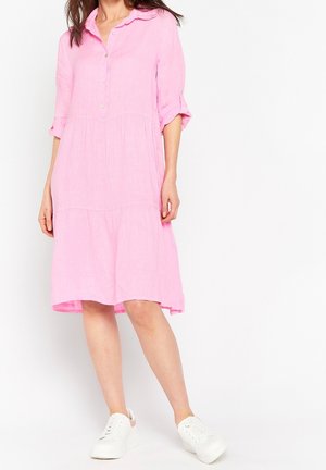 Kobieta w luźnej, różowej sukience do kolan z podwiniętymi rękawami i białymi trampkami, stojąca na białym tle.