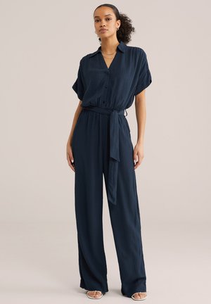 Naine kannab tumesinist tekstuuriga kombekat, millel on lühikesed varrukad, ees nööbitav, vöökohas pael, laiad püksisääred ja valged avatud varbaga kontsad.