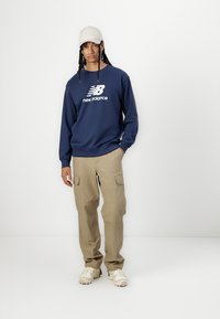 Tumši zila sporta krekla ar baltu logo, bēšīgas bikses ar kabatām un bēšīgas kedas. Modelis valkā gaiša toņa cepuri.