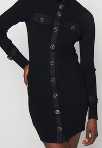 Svart ribbad klänning med långa ärmar, som har två framfickor, guldknappdetaljer och en smal passform, som slutar vid mitten av låret.