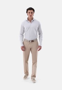 Weißes Langarmhemd, beige Hose, gemusterter Gürtel, beige Schuhe. Das Model steht vor einem schlichten Hintergrund und präsentiert ein lässiges Outfit.