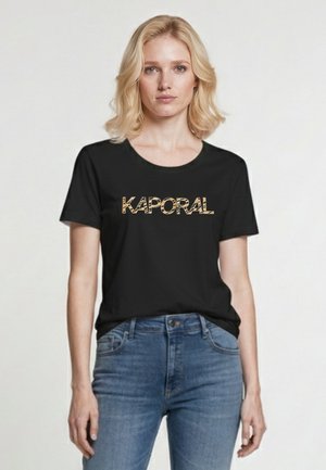 Blondynka w czarnej koszulce Kaporal i niebieskich dżinsach, stojąca na jasnym, jednolitym tle.