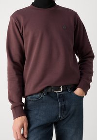 Sudadera burdeos con puños acanalados y cuello redondo, que presenta un pequeño logo en el pecho, combinada con pantalones de mezclilla oscuros y un cinturón negro.