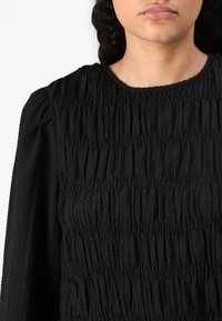Black top with textured, smocked fabric. Features a round neckline and puffed sleeves, showcasing a crinkled surface pattern.