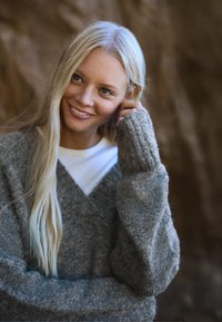 Szary dzianinowy sweter z dekoltem w serek i oversize'owymi rękawami, noszony na białej koszuli. Miękka tekstura, na tle naturalnego, skalistego krajobrazu.