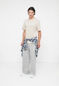 Beige t-shirt med sort tekst, grå sweatpants og en blåternet flannel skjorte bundet om taljen, båret med hvide sko.