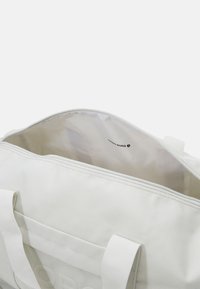 Biała torba sportowa z gładką powierzchnią i kontrastującymi szwami. Posiada dwa uchwyty oraz wewnętrzną metkę z logo, co wskazuje na przestronne wnętrze.