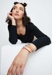 Trois bracelets tressés en rouge, bleu et orange avec des fermoirs en argent reposant sur une main. Le fond est uni et clair, mettant en valeur les accessoires.