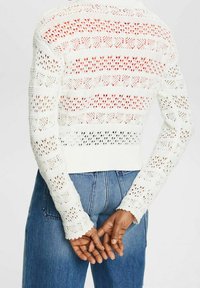 Weißer, gestrickter, verkürzter Pullover mit einem offenen Muster und roten Streifen. Lange Ärmel mit gerippten Bündchen. Kombiniert mit blauen, hochgeschnittenen Jeans.