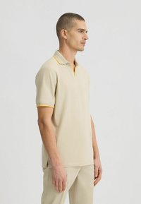 Beige Poloshirt mit strukturiertem Kragen und gelben Akzenten. Kurzärmlig und mit lockerer Passform. Dezentes Logo auf der Brust. Glattes Material.