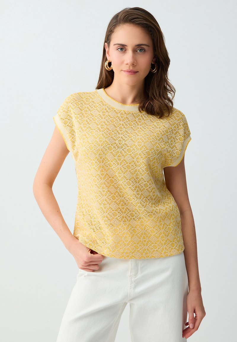 Femme aux cheveux bruns portant des boucles d'oreilles créoles dorées et un haut à manches courtes jaune à motifs, assorti à un pantalon blanc, debout devant un fond uni.