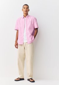 Camicia rosa chiaro a maniche corte con bottoni e colletto classico, abbinata a pantaloni beige e infradito nere. Design semplice, vestibilità casual.