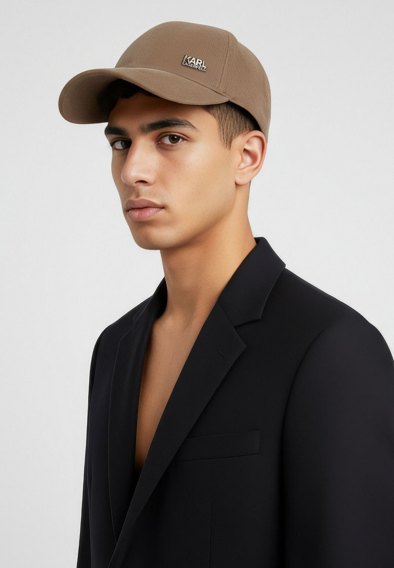 Cappello marrone con design strutturato e dettaglio logo sul davanti, abbinato a un blazer nero, creando un contrasto di colori e texture.